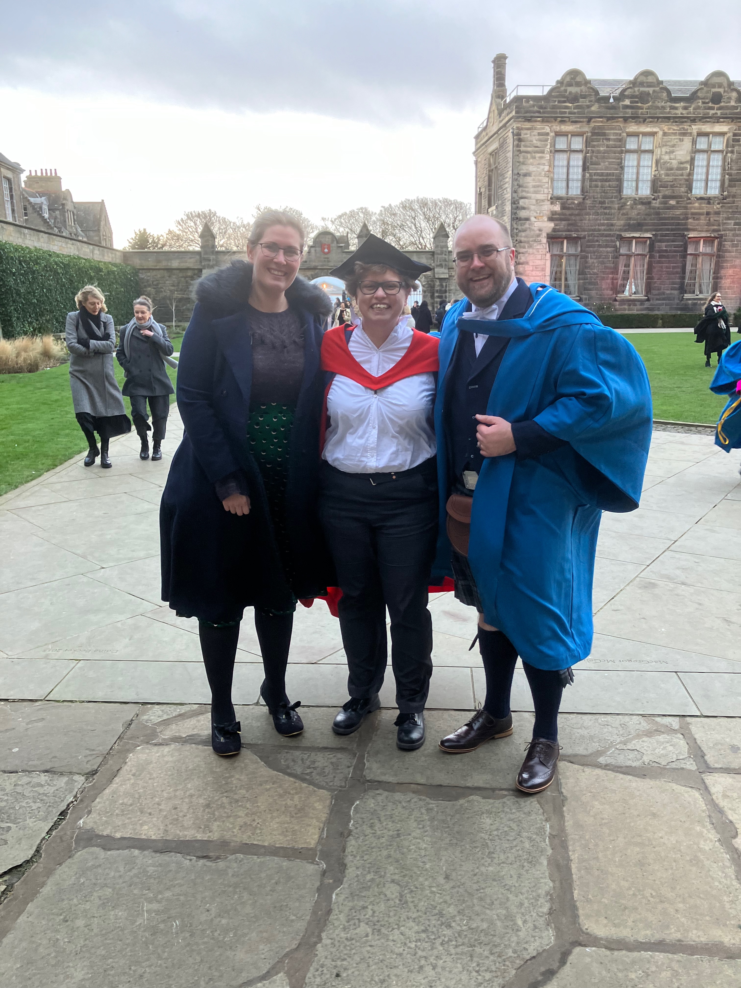 Ronan, Janet and Ronan's wife standing and smiling at the University of St Andrews. Ronan is wearing a kilt and graduation gown.